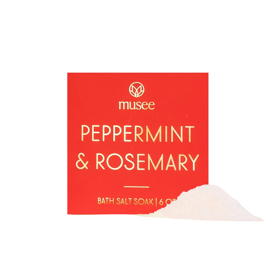 A red square package for musee Peppermint & Rosemary bath salt soak with gold lettering, shown on a white background with a small pile of pale pinkish-white bath salt in front.