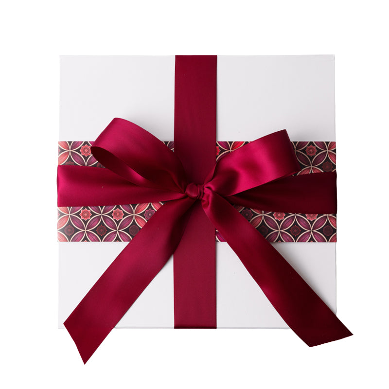 A square white gift box wrapped with a wide burgundy satin ribbon and bow, with a decorative patterned paper strip visible along the sides, on a plain white background.