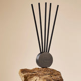 A diffuser with a round dark gray base and five tall black scene sticks, displayed on a textured brown stone against a warm beige background.