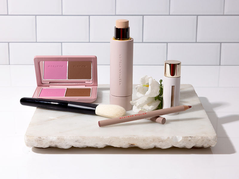 A curated arrangement of Westman Atelier cosmetics on a white marble tray, featuring a pink makeup compact with dual pans, a tall nude-colored foundation stick, a small white and gold bottle, a pencil, and a makeup brush, set against a white subway tile background.