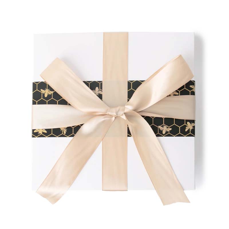 A white gift box with a gold and black pattern featuring bees, adorned with a gold satin bow.